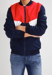 Chaqueta bomber azul marino con diseño de yugo rojo y blanco, cierre de cremallera, puños y dobladillo acanalados, llevada sobre unos vaqueros de mezclilla.