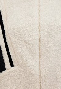 Cremfarbener Fleece-Stoff mit einer weichen, strukturierten Oberfläche. Verfügt über eine zentrale Naht und schwarze Streifenakzente auf einer Seite.