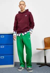 Burgundy hoodie med framficka, gröna sweatpants med vita sidosträck, lager på lager-rutig skjorta och grå sneakers. Enkelt design och material.