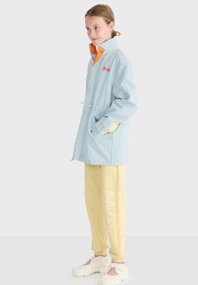 Jeune personne portant une veste bleu clair avec une doublure orange, un pantalon de survêtement jaune avec du texte blanc, et des chaussures blanches épaisses, debout les mains dans les poches.