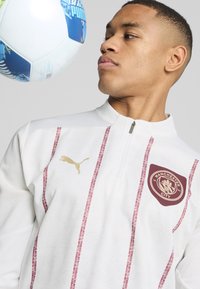 Balta zip-up treniruoklių striukė su raudonomis juostelėmis, auksiniu Puma logotipu ir Mančesterio City emblemą. Yra tekstūruotas audinys ir stovinti apykaklė.