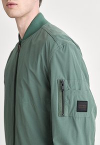 Zelená bomber bunda s vroubkovaným límcem, zipovou kapsou na rukávu a kontrastní látkovou nášivkou na levém rukávu. Hladká textura.