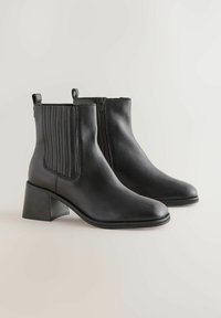 Botine negre din piele, cu vârful ascuțit, detalii subtile de cusătură, panouri laterale elastice și un toc mic de tip bloc. Închidere cu fermoar la spate.
