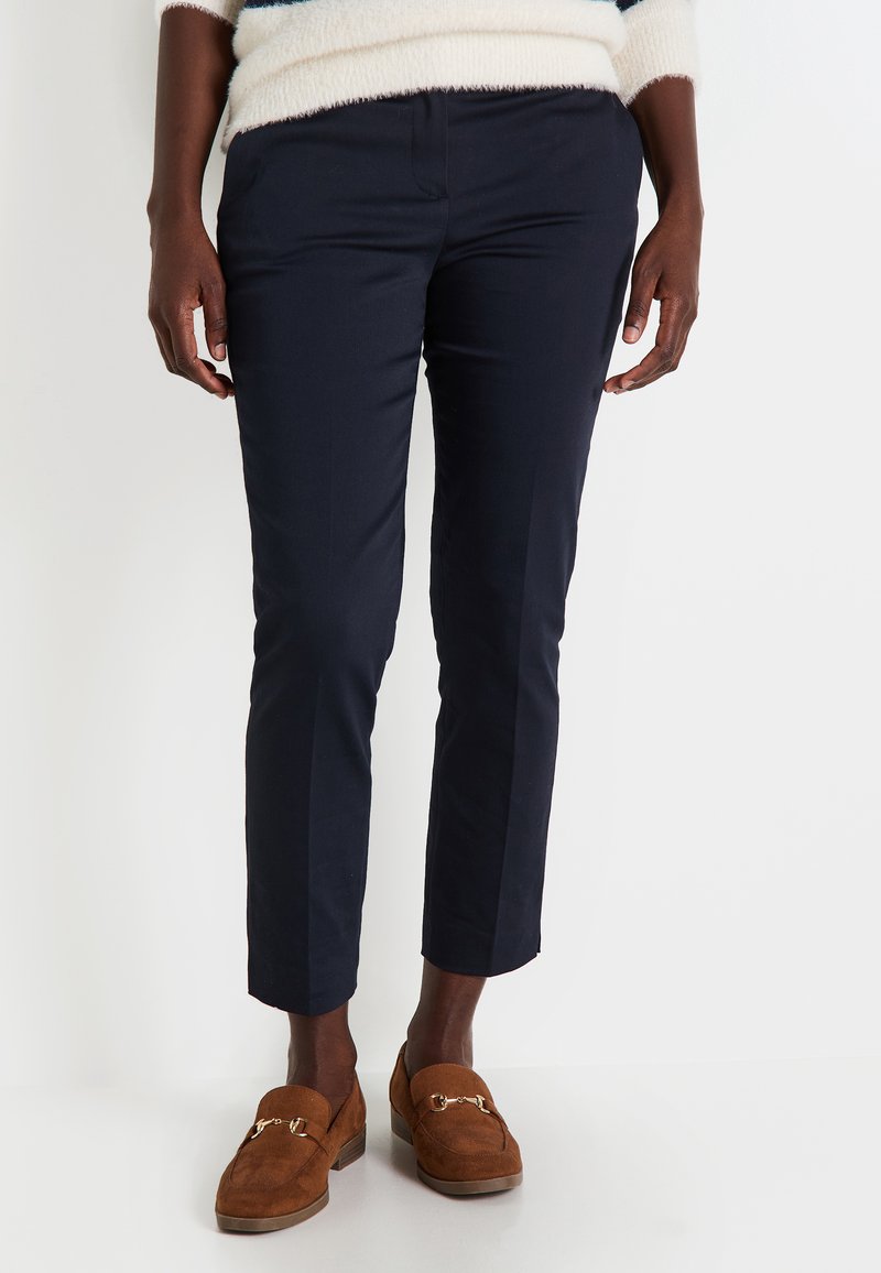 Pantalon ajusté bleu marine, avec une coupe slim, présentant une texture lisse et des plis à l'avant. Porté avec des mocassins en daim marron.