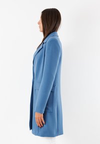 Cappotto azzurro chiaro su misura con rever a punta, tasche laterali e una silhouette lunga e aderente; realizzato in tessuto liscio e testurizzato.