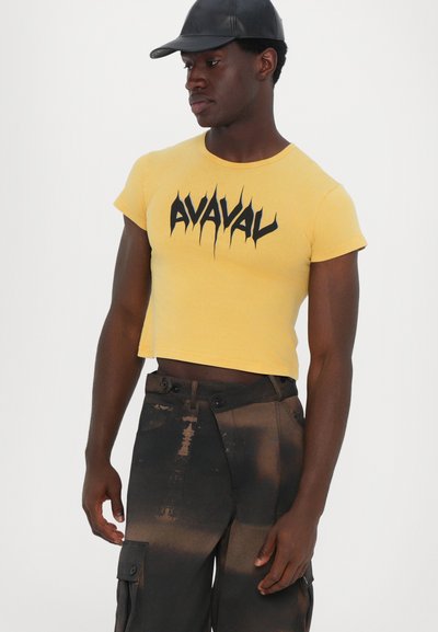 Gul kortærmet t-shirt med sort, aggressiv tekst "AVAVAL." Modellen bærer mørke, strukturerede cargo-bukser med lommer og et falmet mønster.