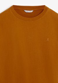 Tricou de culoare ruginită cu guler rotund, având un mic logo brodat cu craniu și oase încrucișate pe partea stângă a pieptului și o etichetă Scalpers vizibilă în interiorul gulerului.