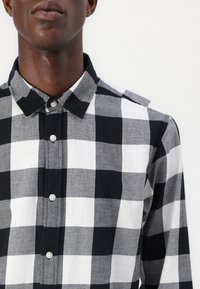 Camicia button-up a scacchi bianca e nera con colletto, caratterizzata da una texture morbida e quadrati a contrasto di varie dimensioni.