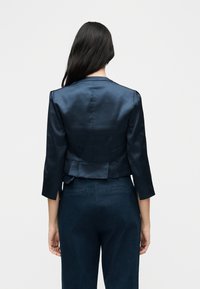 Femme aux longs cheveux foncés portant un blazer en satin bleu marine à manches trois-quarts et un pantalon bleu marine, vue de dos sur un fond uni.