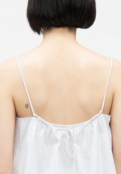 Άτομο με κοντά σκούρα μαλλιά, φοράει λευκό τοπ με λεπτές ρίγες και λεπτές τιράντες, που δείχνει το πάνω μέρος της πλάτης και ένα μικρό τατουάζ στην αριστερή ωμοπλάτη.