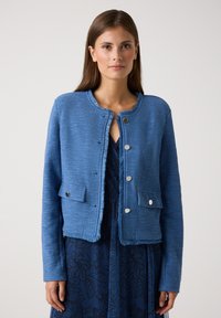 Veste courte bleu texturée à manches longues, col rond et boutons-pression argentés. Deux poches avant améliorent la fonctionnalité.