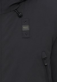 Manteau isolant noir avec une poche zippée, des détails en bouton et une patch de marque. Le matériau est lisse et légèrement texturé.
