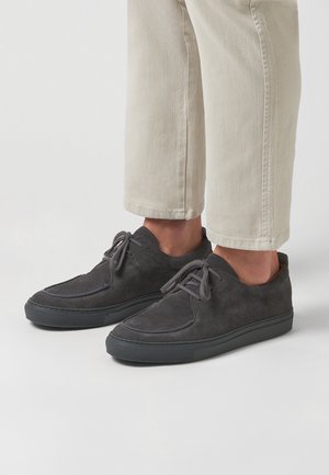 Zapatos de ante gris oscuro con puntera redondeada, costuras mínimas, diseño de cordones y suela de goma negra texturizada. Estilo informal con líneas limpias.