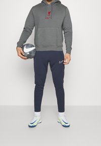 Sweat à capuche gris avec logo Liverpool rouge, pantalon de jogging bleu marine, chaussettes blanches et crampons de football colorés ; tenant un ballon de football gris et blanc.