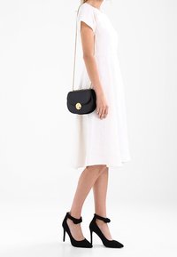 Musta nahkainen olkalaukku, jossa kultainen pyöreä lukko. Malli käyttää polvipituista valkoista pellavamekkoa ja mustia mokkanahkaisia korkeakorkoisia kenkiä.