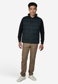 Gepolsterte marineblaue Weste mit hohem Kragen und Vorderreißverschluss, kombiniert mit beigen Hosen und weißen Sneakers, die einen legeren, geschichteten Look erzeugen.