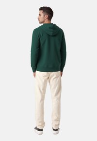 Mørkegrøn hættetrøje med snor i hætten, ribbede manchetter og kant; parret med lysebeige bukser og sorte sneakers.