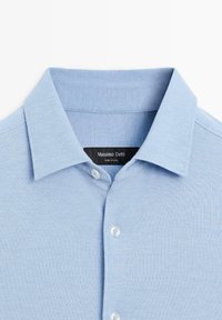 Jasnoniebieska koszula z guzikami, wykonana z teksturowanej tkaniny, z rozpiętym kołnierzykiem i białymi guzikami. Metka informuje "Massimo Dutti, wyprodukowano w Chinach."