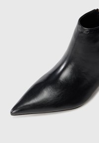 Bottine en cuir noir avec un bout pointu et une finition lisse et brillante sur un fond gris clair uni.