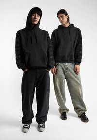Felpe con cappuccio nere e maniche lunghe con grafiche stampate. Un modello indossa pantaloni scuri, l'altro in jeans chiari strappati.
