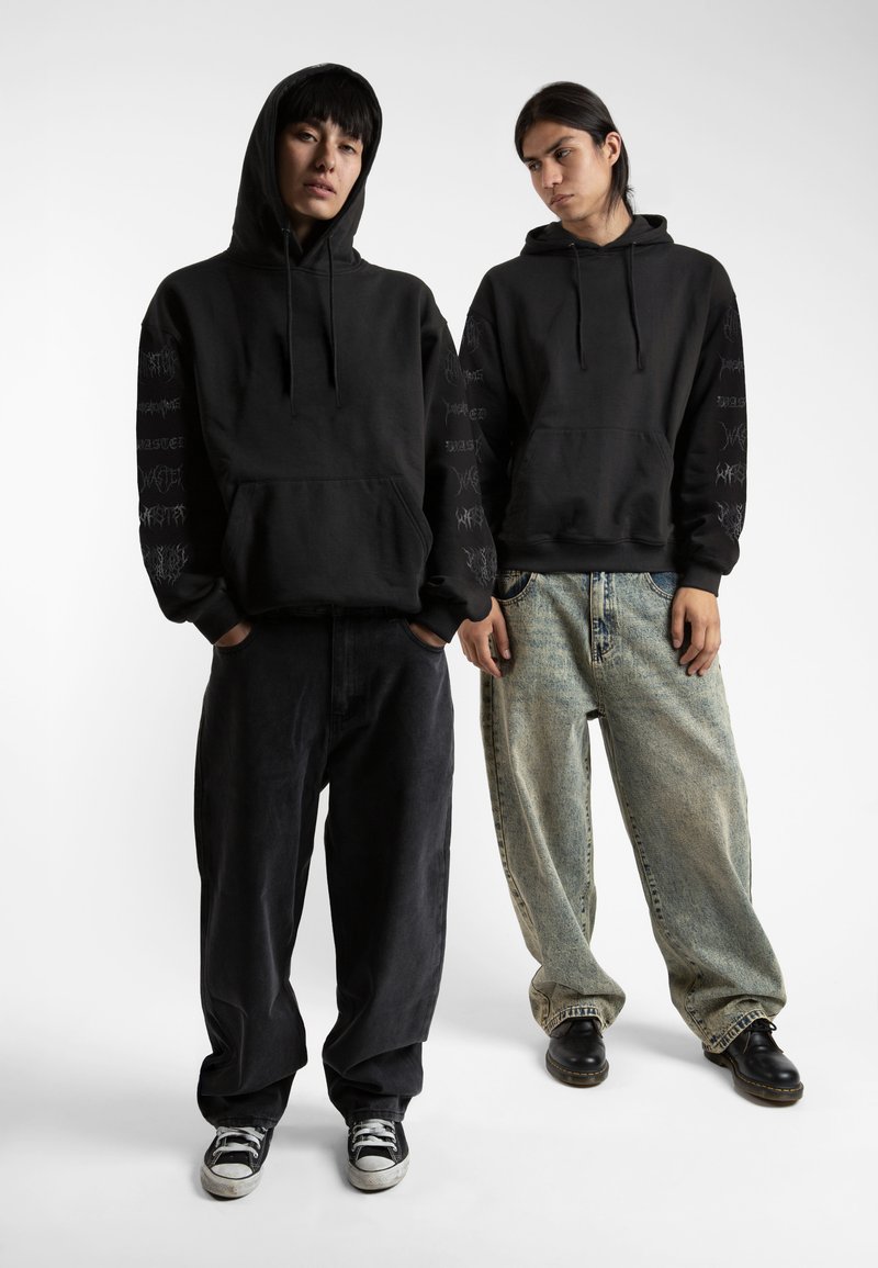 Felpe con cappuccio nere e maniche lunghe con grafiche stampate. Un modello indossa pantaloni scuri, l'altro in jeans chiari strappati.