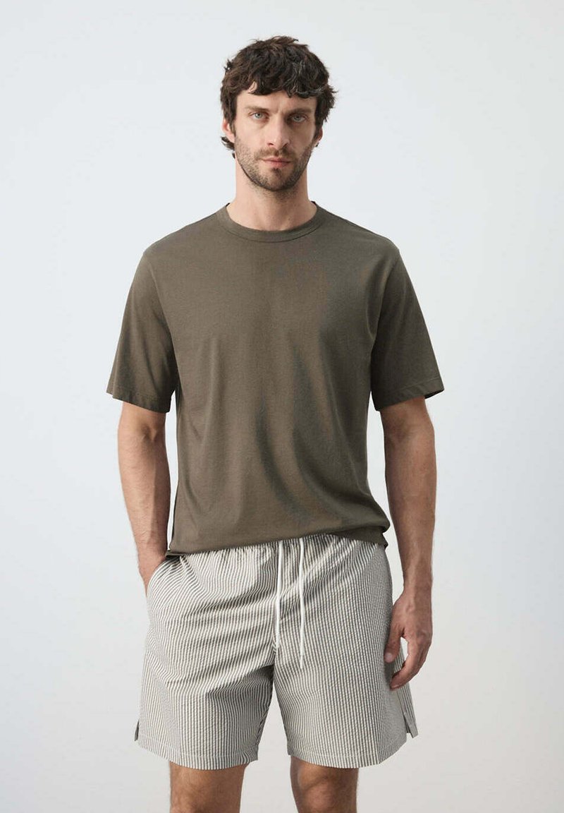 Homme aux cheveux courts et bouclés portant une chemise à manches courtes vert olive et un short clair rayé avec cordon, debout devant un fond uni.