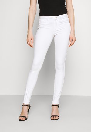 Vrouw die witte skinny jeans, een zwart mouwloos topje en zwarte hoge hak sandalen met enkelbandje draagt, staand tegen een effen achtergrond.