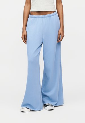 Osoba stojaca pred jednofarebnym pozadim, oblecena v svetlomodrych nohaviciach s sirokymi nohavicami a elastickym pasom, v ciernych a bielych teniskach.