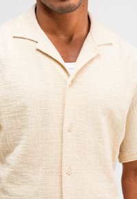 Beige shirt met korte mouwen, gemaakt van gestructureerde stof, met een knoopsluiting aan de voorkant en een kraag, en subtiele patronen door het geheel.