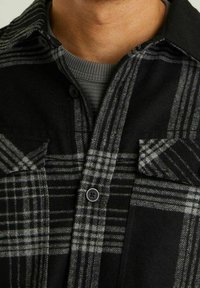 Camicia in flanella a quadratini neri e grigi con bottoni, due tasche sul petto e una consistenza morbida. Il tessuto presenta modelli a scacchi alternati.