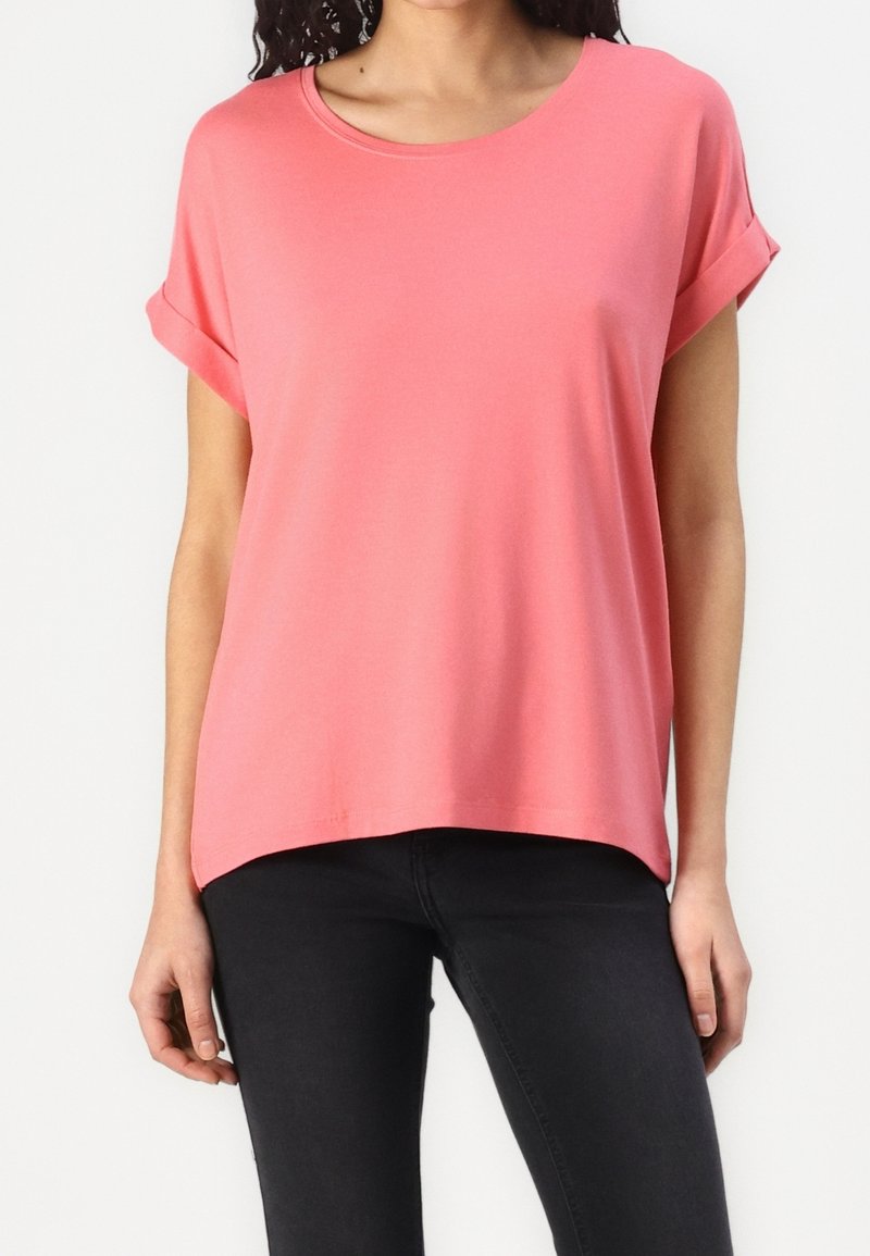 Femme portant un haut ample rose corail à manches courtes avec revers et un pantalon noir slim, debout devant un fond blanc uni.