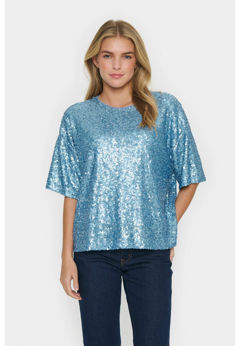 Ljust blå paljettblus med en lös passform och korta ärmar. Den texturerade ytan reflekterar ljus och förstärker den livliga färgen. Matchad med mörka jeans.