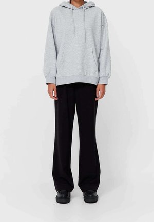 Osoba oblečená v světle šedé mikině s kapucí a přední kapsou, černé široké kalhoty a černé boty na platformě, stojící před jednobarevným pozadím.