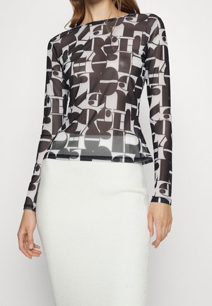 Femme portant un haut à manches longues à motif abstrait noir et blanc avec une jupe blanche ajustée, debout les bras détendus le long du corps.