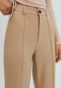 Be�żowe dopasowane spodnie o gładkiej teksturze, z przodem z zakładkami oraz zapięciem na guzik w talii. Ręka trzymająca pas z boku.