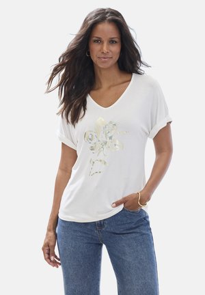 Frau mit langen dunklen Haaren trägt ein weißes V-Ausschnitt-T-Shirt mit goldener Blume und dem Text „Best Day Ever“, kombiniert mit blauen Jeans und goldarmband.