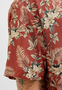 Camisa de manga corta en terracota con patrones florales y de hojas en beige. Hecha de una tela ligera con un corte holgado y una textura suave.