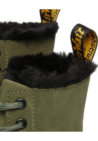 Close-up van olijfgroene laarzen met zwarte bontvoering en geel-zwarte merk trekbanden, die metalen veterogen en grijze veters tonen.