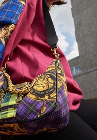 Borsa a scacchi viola con accenti floreali dorati, una tracolla a catena e una toppa verde a contrasto, progettata da Versace Jeans Couture.