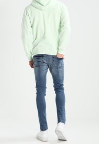 Persona che indossa un hoodie verde chiaro, jeans slim-fit blu e scarpe da ginnastica bianche, in piedi con una mano nella tasca posteriore, rivolta di spalle.