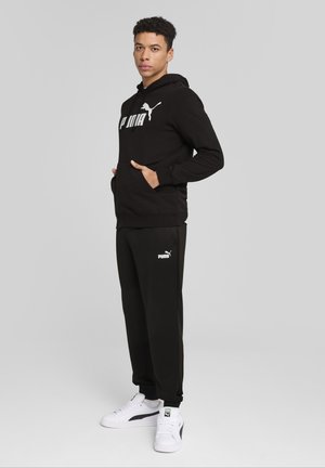 Sort hættejakke med hvid "PUMA" logo, parret med matchende sorte joggers med PUMA logoet. Båret med hvide sportsko.