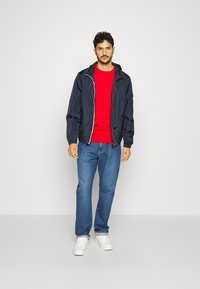 Giacca a vento blu navy con cappuccio, chiusura con zip e polsini elasticizzati sopra un maglione rosso. Jeans blu e sneaker bianche completano l'outfit.