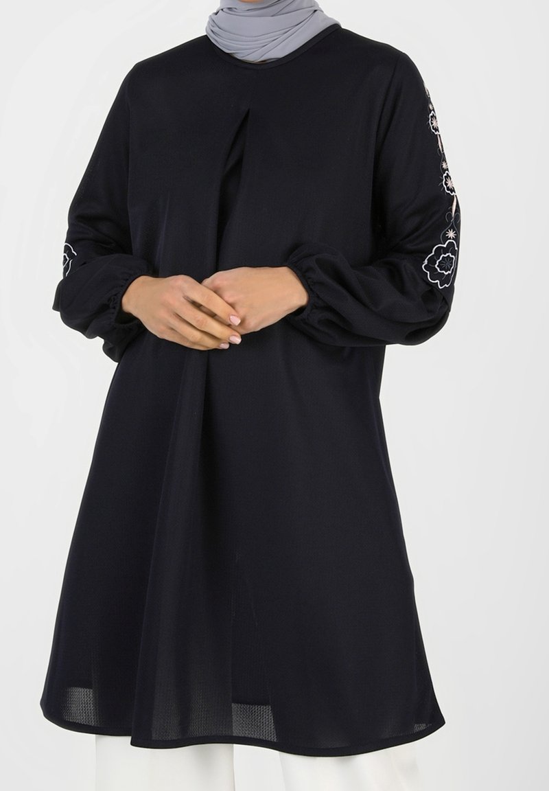 Femme portant une longue tunique noire avec des broderies florales sur les manches et un hijab gris clair, les mains jointes à la taille.