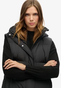  Schwarze gepolsterte Weste mit Hochkragen, Druckknöpfen und Reißverschluss vorne. Getragen über einem strukturierten dunklen Pullover. Das Modell hat die Arme verschränkt.