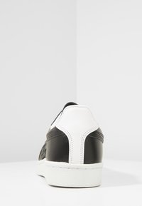 Sneaker bianco e nero con finitura in pelle verniciata, accenti laterali testurizzati e suola in gomma bianca. Presenta curve morbide e dettagli di cucitura.