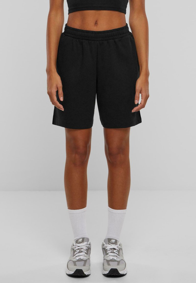 Shorts negri din bumbac cu talie elastică, până la genunchi, asociați cu șosete albe cu nervuri și pantofi sport gri deschis.
