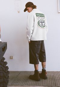 Giacca in pile grigia con logo Toyota Land Cruiser, pantaloncini neri e stivali marrone scuro. In piedi su pavimento strutturato con sfondo a parete.