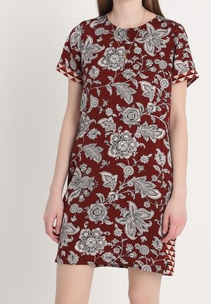 Kvinde iført kortærmet, over knæet vinrød kjole med hvidt blomstermønster og geometrisk design på indvendige ærmekanter og sideslidser.