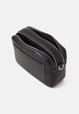 Trousse de toilette en cuir noir avec deux compartiments zippés ouverts et une poche extérieure avec des coutures blanches sur un fond blanc.