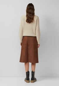 Femme aux longs cheveux bruns portant un pull en tricot beige, une jupe marron longueur genou et des bottines noires, se tenant devant un fond gris uni.